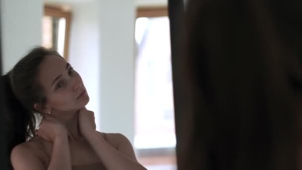 Молодая брюнетка в бежевом топе вытягивает шею перед зеркалом — стоковое видео