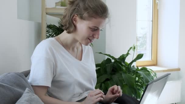 Улыбающаяся молодая женщина, сидящая на диване, используя ноутбук, набирая сообщение — стоковое видео