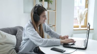 Kulaklıklı neşeli bayan öğrenci evde dizüstü bilgisayar kullanıyor, müzik dinliyor.