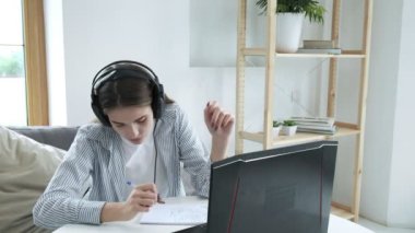 Kulaklıklı neşeli bayan öğrenci evde dizüstü bilgisayar kullanıyor, müzik dinliyor.