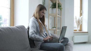 Gülümseyen genç kadın kanepede oturuyor, dizüstü bilgisayar kullanıyor, mesaj yazıyor.