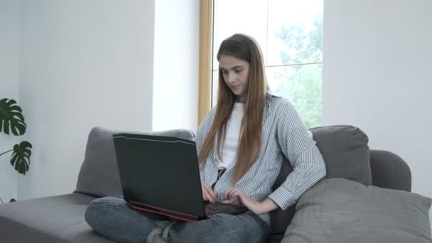 Glimlachende jonge vrouw zitten op de bank, met behulp van laptop notebook, typen bericht — Stockvideo
