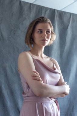 Kısa saçlı, pembe elbiseli beyaz bir kadın stüdyoda kollarını kavuşturmuş poz veriyor.