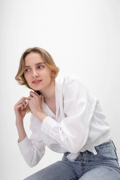 Молода мила усміхнена біла дівчина позує в білій сорочці, блакитні джинси в студії — стокове фото