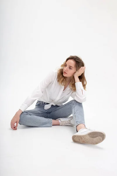 Caucasian woman posing in shirt and blue jeans, sitting on white studio floor — Stock Photo, Image