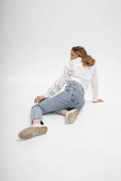 Beyaz kadın gömlek ve kot pantolon giymiş, beyaz stüdyo zemininde oturuyor. — Stok fotoğraf