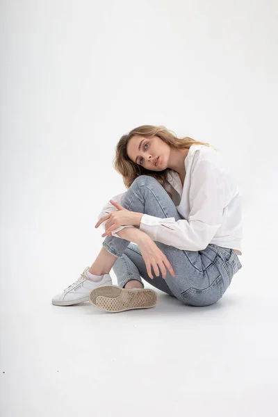 Caucasian woman posing in shirt and blue jeans, sitting on white studio floor — Stock Photo, Image