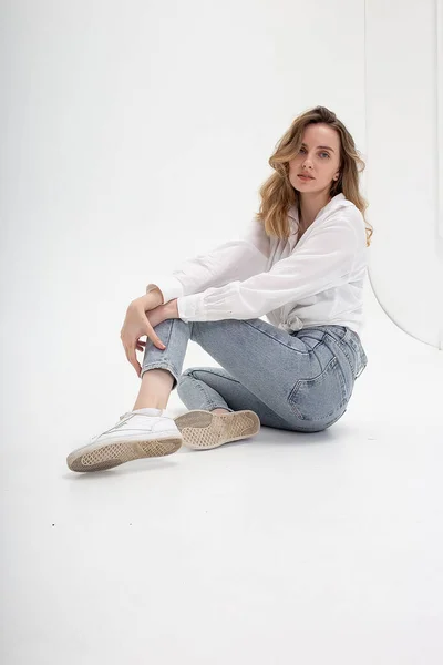 Mujer caucásica posando en camisa y jeans azules, sentado en el piso del estudio blanco — Foto de Stock