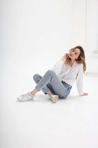 Kaukasiska kvinna poserar i skjorta och blå jeans, sitter på vit studio golv — Stockfoto