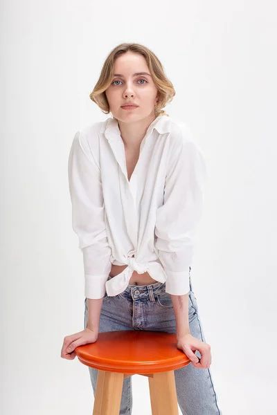 Young caucasian girl in white shirt, blue jeans standing near chair at studio — Stock Photo, Image