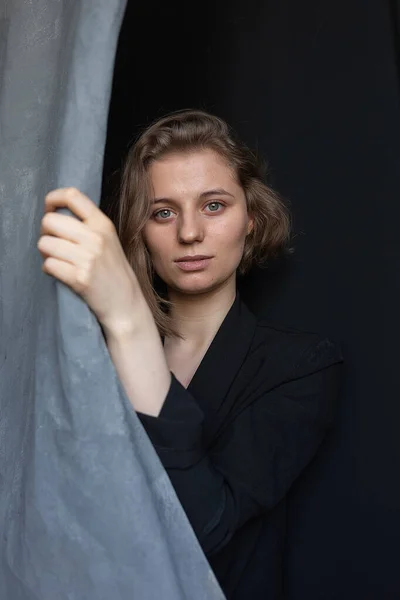 caucasian woman with short hair posing in black suit jacket, holding curtain
