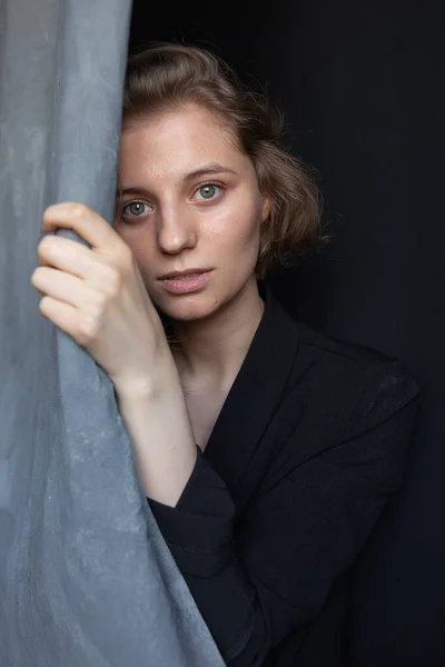 caucasian woman with short hair posing in black suit jacket, holding curtain