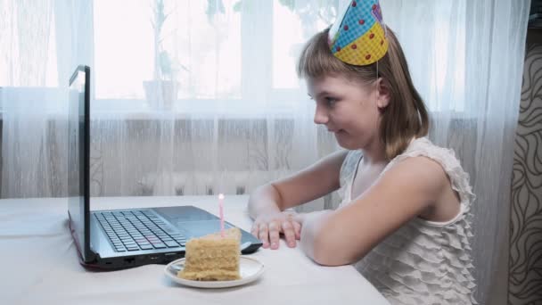 Chica se sienta a la mesa con el ordenador portátil, sopla vela encendida en pastel de cumpleaños — Vídeos de Stock