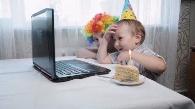 Çocuklar dizüstü bilgisayarla masaya oturur, doğum günü pastasının üzerine mum üflerler.