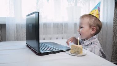 Çocuk dizüstü bilgisayarla masada oturuyor, doğum günü pastasına mum üflüyor.