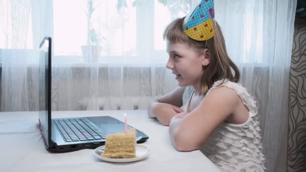 Meisje zit aan tafel met laptop computer, blaast aangestoken kaars op verjaardag taart — Stockvideo