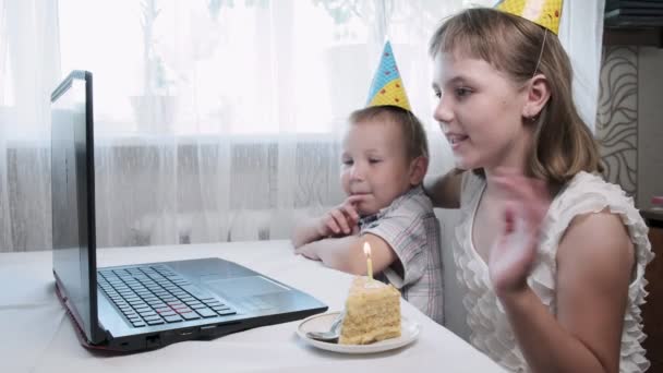 Дети сидят за столом с ноутбуком, задувают свечи на торте день рождения — стоковое видео