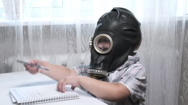 Kleine jongen met gasmasker zit aan tafel, tekent krabbels met pen in notitieboekje — Stockvideo