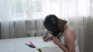Gaz maskeli kız masada oturuyor, resimlerini çiziyor albümde keçeli kalemlerle.
