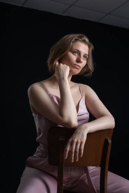Pembe takım elbiseli kısa saçlı kadın, siyah arka planda sandalyede oturuyor.