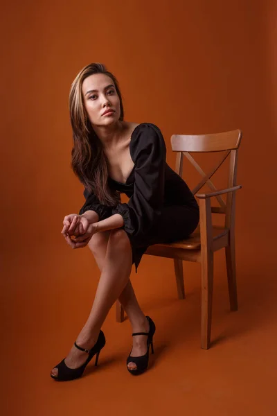 pretty model in black dress, highheel shoes sits in chair on orange background