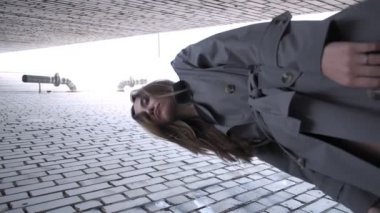 Asyalı güzel bir model. Trençkot giyiyor. Alt noktadan çekilen duvarların yanında duruyor.