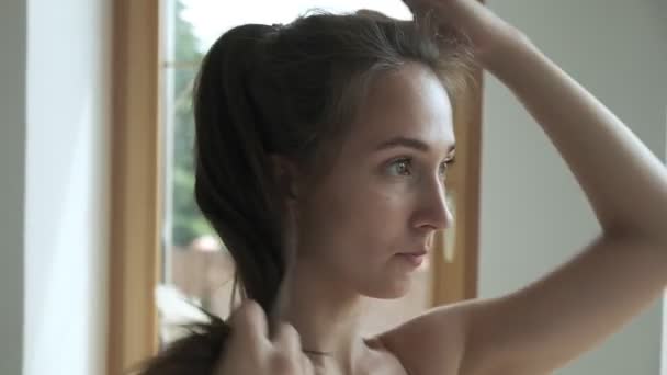 Sporty brunette pretty female stand at window, fixes hair, collects hair in bun — Stok Video