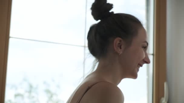 Hübsche brünette Frau mit gesammelten Haaren steht am Fenster, lächelt und lacht — Stockvideo
