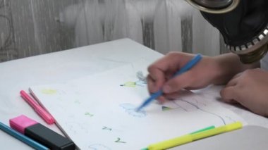 Gaz maskeli kız masada oturuyor, resimlerini çiziyor albümde keçeli kalemlerle.