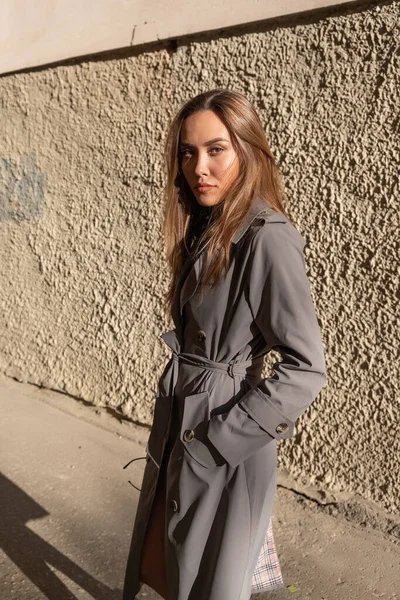 야외 벽 가까이에 서 있는 회색 도랑 코트를 입고 있는 매력적 인 젊은 아시아인 모델 — 스톡 사진