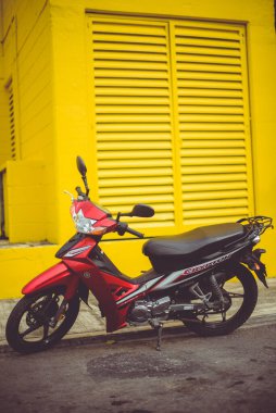 Kırmızı motosiklet arka planda parlak sarı binayla caddeye park edilmiş.