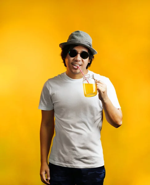 亚洲男子喝橙汁戴墨镜和软呢帽帽子反对黄色背景 — 图库照片