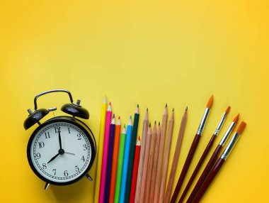 Çalar saat, renkli kalemler ve sarı zemin üzerine fırça