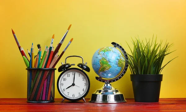 World Globe, Alarm Clock, Plant and School Stationary in Basket on Wooden Table Yellow Background