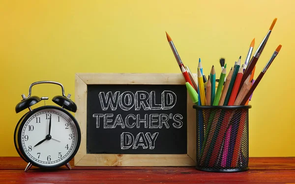 World Teacher\'s Day Text. Alarm Clock, Blackboard and School Stationary in Basket on Wooden Table Yellow Background