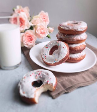 Beyaz tabakta beyaz kremalı ev yapımı çörekler ve bir bardak süt ve gri arka planda pembe güller.