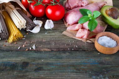 Bir demet İtalyan spagettisi, çiğ soba eriştesi, çiğ tavuk eti, ahşap arka planda domatesli avokado, fotokopi alanı ile derisi yüzülmüş. Ev yapımı yemek pişirme kavramı