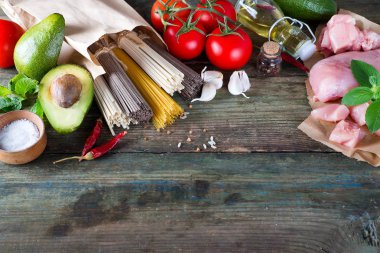 Bir demet İtalyan spagettisi, çiğ soba eriştesi, çiğ tavuk eti, ahşap arka planda domatesli avokado, fotokopi alanı ile derisi yüzülmüş. Ev yapımı yemek pişirme kavramı