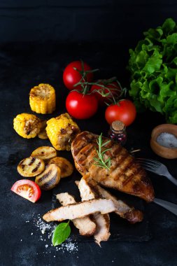 Çam dilimi ve tavuk ızgara biftek, domates, mısır ve patates ızgarada fesleğen ve biberiye ile siyah beton arka planda.