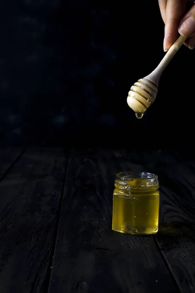 A menina está segurando uma colher de madeira em suas mãos, em mel líquido vara em frasco de mel no fundo preto — Fotografia de Stock
