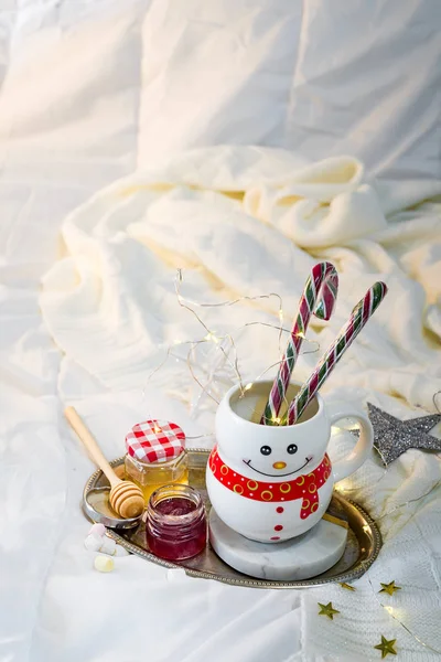 Cozy soft blanket with a cup in the form of a snowman with Christmas candies, honey and jam on the bed with flashlights. Cristmas concept lazy morning, warm winter mood — Stock Photo, Image