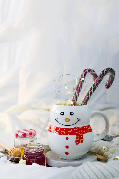 Уютное мягкое одеяло с чашкой в виде снеговика с рождественскими конфетками, медом и джемом на кровати с фонариками. Рождественская концепция ленивое утро, теплое зимнее настроение — стоковое фото