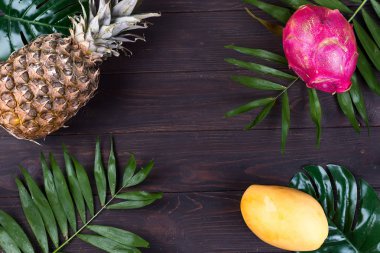 Koyu ahşap arka plan üzerinde düz yatıyordu tropikal meyve çerçeve ile ananas, dragon meyve, sarı mango ve palmiye yaprakları