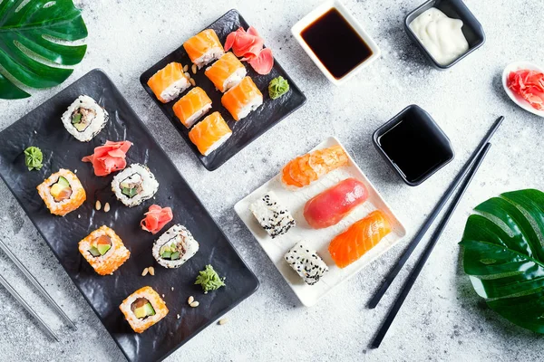 Varios sushi fresco y delicioso establecido en pizarra blanca y negra con palos de metal y pizarra, salsa y hojas verdes sobre fondo de piedra clara, plano laico — Foto de Stock