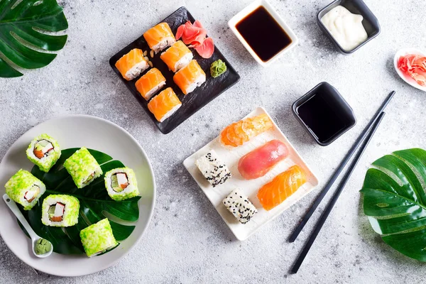 Varios sushi fresco y delicioso establecido en pizarra blanca y negra con palos de metal y pizarra, salsa y hojas verdes sobre fondo de piedra clara, plano laico — Foto de Stock