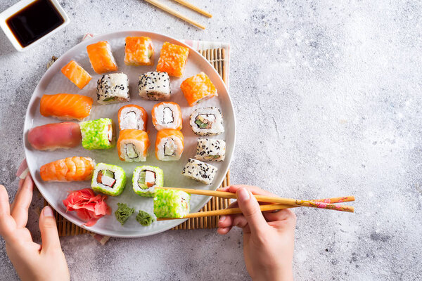 Разнообразные свежие и вкусные суши на белой доске, рука с деревянными палочками, соус на светлом каменном фоне, плоский уголок
