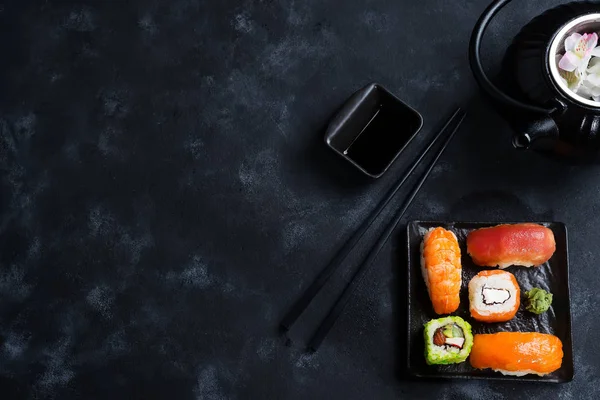 Asian food background with black iron teapot and sushi set on slate plate on black stone table, flat lay
