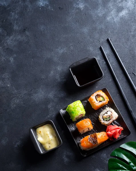 Varios sushi fresco y delicioso establecido en pizarra negra con palos de pizarra, salsa y nori sobre fondo de piedra negra, plano laico — Foto de Stock