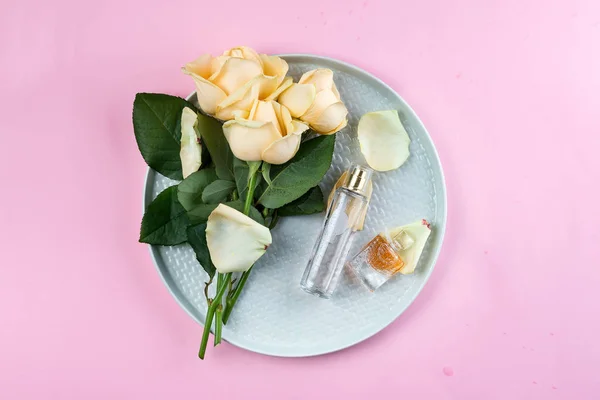 Butelki perfum otoczone kwiat róży na talerzu na różowym tle, płaskie lay — Zdjęcie stockowe