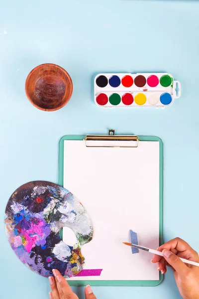 Clipboard with watercolor paints in box, in the hands of brushes and palette on blue table. Top view. Copy space — Stock Photo, Image